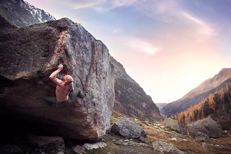 Een man zonder shirt is aan het boulderen op een grote rots in een bergachtig landschap bij zonsondergang; onder een kleurrijke hemel zijn dennenbomen en bergtoppen in de verte te zien.